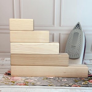 Può includere: Un set di sei blocchi di legno rettangolari in varie tonalità di marrone chiaro, disposti a gradini. Un ferro da stiro Philips grigio e bianco è posizionato a destra. I blocchi poggiano su un tessuto a fantasia floreale.