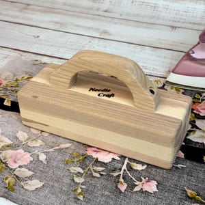 Può includere: Un blocco per lavori di cucito in legno con manico. Il blocco è realizzato in legno chiaro con un manico e le parole "Needle Craft" stampate sul lato. Il blocco è rettangolare e ha una finitura liscia.