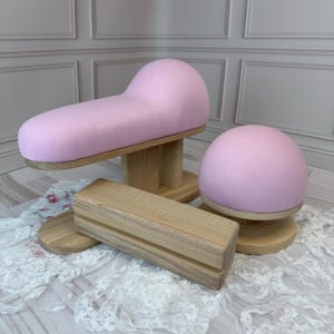 Pode incluir: Um conjunto de três ferramentas de alfaiataria em madeira com superfícies cobertas por tecido rosa claro. A coleção inclui uma almofada de passar longa e curva, uma almofada em forma de cúpula e um bloco de madeira com acabamento natural.