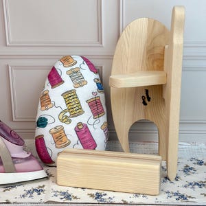 May include: A wooden pressing ham, a wooden sleeve board, and a small wooden block are displayed. A white ironing board cover with a colorful spool design is also present. A pink iron is visible on the left.