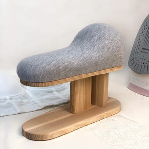 May include: A gray padded sleeve board with a wooden base and supports. The board is designed for ironing sleeves and other small garment areas. A gray iron is visible in the background.