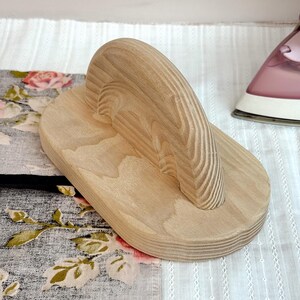 Può includere: Un battente in legno chiaro, utilizzato per stirare le cuciture nel cucito. Il battente ha una base rettangolare con un manico curvo. Lo sfondo include tessuto floreale e un ferro da stiro rosa.