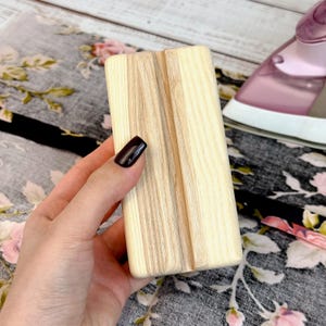 Può includere: Un supporto per telefono rettangolare in legno con un motivo a venature chiare. Il supporto è tenuto in mano. Lo sfondo include un tessuto floreale e un ferro da stiro rosa.