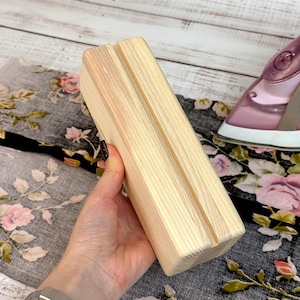 Può includere: Un supporto per telefono in legno chiaro con una scanalatura sulla parte superiore per tenere il telefono. Il supporto è rettangolare e realizzato in legno non rifinito. Lo sfondo include un tessuto a fantasia floreale e un ferro da stiro rosa.