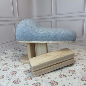 Op de afbeelding: Een kleermakerskussen met een grijs gemêleerde stoffen hoes en een lichtgekleurde houten voet, vergezeld van een apart rechthoekig houten persblok. Het kleermakerskussen heeft een gebogen, ronde vorm voor het persen van naden.