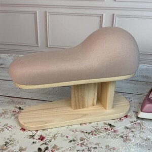 May include: A light pink padded pressing ham on a natural wood base. The ham is oval-shaped and mounted on a wooden stand with a rounded base. The wooden base has a natural finish.