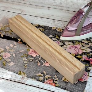 Può includere: Un supporto per asse da stiro in legno chiaro con due scanalature. Il supporto è rettangolare e poggia su un tessuto a fantasia floreale. Un ferro da stiro rosa e grigio è sullo sfondo.