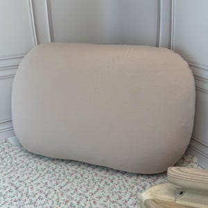 Op de afbeelding: Een groot, beige, langwerpig kussen. Het kussen is gemaakt van een zachte, getextureerde stof. Het kussen ligt op een oppervlak met bloemenpatroon. Het kussen staat in de hoek van een kamer met witte muren.