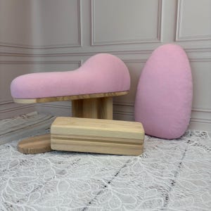 May include: A set of sewing pressing tools including a curved pink fabric-covered tailor's ham, a matching pink oval pressing cushion, and a rectangular wooden clapper with a natural light wood grain finish and smooth rounded edges.