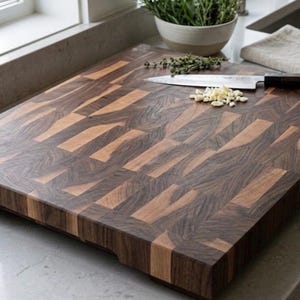 May include: A large, rectangular wooden cutting board with a dark brown and light brown block pattern. A stainless steel knife and chopped garlic are on the board. A small bowl of herbs sits in the background.