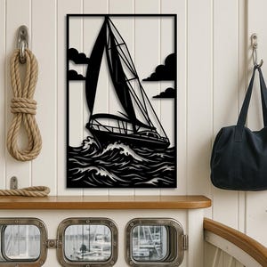 May include: Black metal wall art depicting a sailboat at sea. The sailboat is shown with its sails billowing in the wind, navigating through choppy waves. The artwork includes dark clouds and is framed in a rectangular shape.