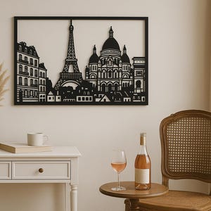 May include: A black metal wall art piece showing the Paris skyline, including the Eiffel Tower and Sacré-Cœur Basilica. The artwork is framed and hangs above a white table with a vase of dried flowers, a book, and a coffee cup.