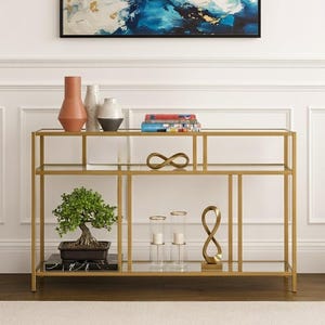 Könnte beinhalten: Eine goldfarbene Konsole mit Glasböden präsentiert dekorative Gegenstände. Ein Bonsai-Baum, Vasen, Bücher und Skulpturen sind auf den Regalen angeordnet. Ein abstraktes Gemälde in Blau-, Weiß- und Goldtönen hängt darüber.
