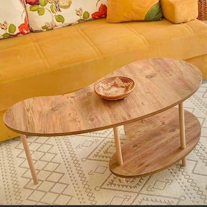 Può includere: Un tavolino in legno a due livelli con una forma unica a fagiolo. Il tavolo ha una finitura a venatura chiara e poggia su quattro gambe cilindriche. Un piccolo cestino intrecciato è posto sul ripiano superiore. Il tavolo è posizionato su un tappeto fantasia.