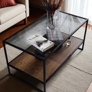 May include: A contemporary coffee table with a clear glass top and a wooden lower shelf. The table has a black metal frame. A book, decorative items, and a vase with dried flowers are on the table. The table sits on a beige rug.
