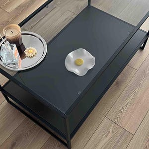 May include: A rectangular coffee table with a black metal frame and two tiers, featuring a clear glass top and a solid black lower shelf. On the glass surface sits a white ceramic dish shaped like a fried egg, alongside candles and a silver tray.