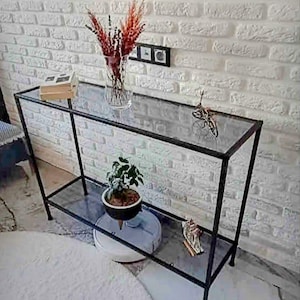 Op de afbeelding: Een moderne console met een zwart metalen frame en twee glazen planken. De bovenste plank bevat een transparante vaas met gedroogde bloemen, boeken en een decoratieve fiets. Een kleine potplant en een decoratief object staan op de onderste plank. De tafel is 120 cm lang.