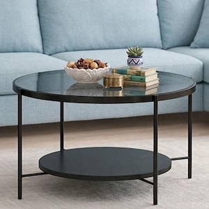 Könnte beinhalten: Ein runder Couchtisch aus schwarzem Metall mit Glasplatte und unterem Regal. Der Tisch steht vor einem hellblauen Sofa. Eine weiße Schale mit Potpourri, eine kleine Sukkulente und Bücher stehen auf dem Tisch.