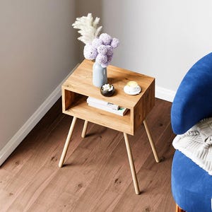 May include: A small, light-colored wooden side table with a rectangular top and angled legs. The table has a lower shelf and holds a vase of purple flowers, a small dish, and a teacup. The table is next to a blue armchair.
