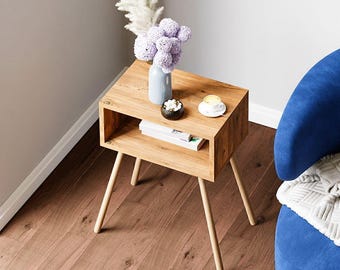 May include: A small, light-coloured wooden side table with a rectangular top and angled legs. The table has a lower shelf and holds a vase of purple flowers, a small dish, and a teacup. The table is next to a blue armchair.