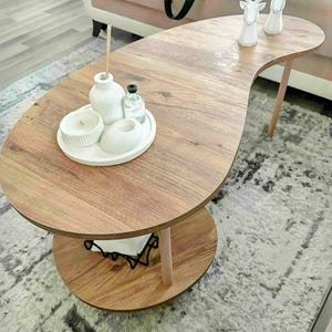 Puede incluir: Una mesa de centro de madera en forma de riñón con un estante inferior. La mesa exhibe una bandeja blanca con una vela, un jarrón y una botella pequeña. Dos figuras de ciervos blancos están sobre la mesa. La mesa está sobre una alfombra estampada.