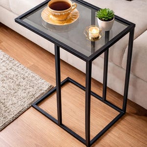 May include: A modern side table with a black metal frame and a clear glass top. A cup of tea on a saucer, a small potted plant, and a candle sit on the table. The table is next to a beige sofa.