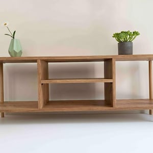 May include: A light brown wooden entertainment center with a geometric vase and a potted plant. The unit has two shelves and a central compartment with two shelves. The unit is 60 inches long, 15 inches deep, and 20 inches high.