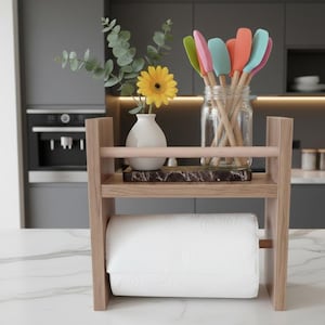 May include: A wooden kitchen organizer with a paper towel roll on the bottom shelf. The top shelf holds a small white vase with a yellow flower, a marble cutting board, and a glass jar filled with colorful spatulas.