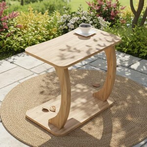 May include: A light-colored wooden side table with a curved leg design. The table has a rectangular top and a lower shelf. A cup of coffee sits on the table, which is placed on a round woven mat. The background features greenery and flowers.
