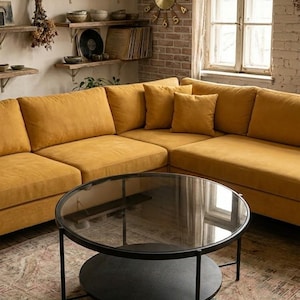 Pode incluir: Uma sala de estar moderna com um grande sofá seccional amarelo mostarda e uma mesa de centro redonda com estrutura de metal preta e tampo de vidro. Um relógio dourado está pendurado em uma parede de tijolos e prateleiras de madeira exibem decoração.