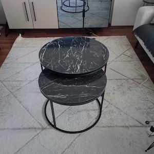 May include: Two-tiered, round side tables with black marble-look tops and black metal frames. The tables are nested, with the smaller table positioned beneath the larger one. The tables are on a light-colored rug.