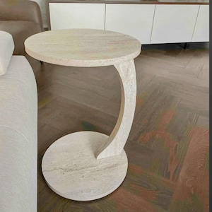 Può includere: Un tavolino rotondo beige chiaro con un supporto curvo e una base circolare. Il piano del tavolo ha un diametro di circa 50 cm. Il design è minimalista e moderno, adatto per un soggiorno o un salotto.