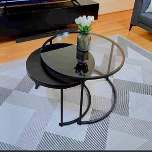 Op de afbeelding: Een moderne salontafel met twee niveaus, met een zwart metalen frame en een helder glazen blad. De onderste laag is effen zwart. Een vaas met witte bloemen staat op de bovenste laag. De tafel staat op een patroonkleed.