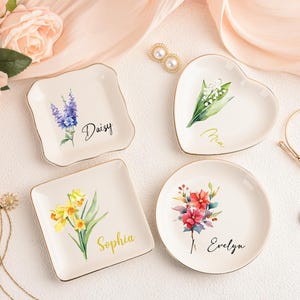Könnte beinhalten: Vier dekorative Keramik-Schmuckschalen mit floralen Designs und goldenen Rändern. Die Schalen haben verschiedene Formen: quadratisch, herzförmig und rund. Jede Schale trägt einen Namen: Daisy, Mia, Sophia und Evelyn. Auch Schmuck und Blumen sind vorhanden.