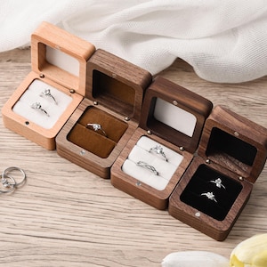 May include: A collection of wooden ring boxes, each open to display a ring. The boxes vary in color from light to dark wood, with velvet or fabric interiors in white, brown, and black. The rings are silver with clear stones.