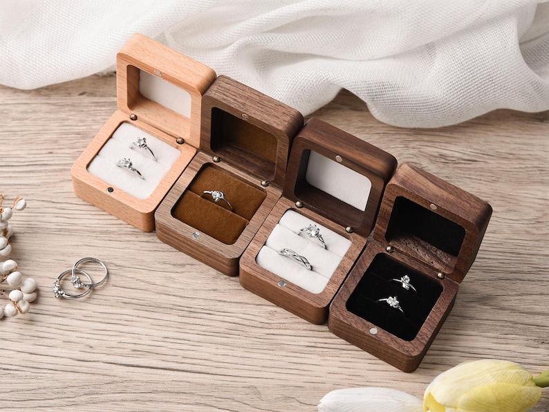 May include: A collection of wooden ring boxes, each open to display a ring. The boxes vary in color from light to dark wood, with velvet or fabric interiors in white, brown, and black. The rings are silver with clear stones.