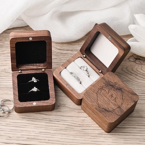 May include: Wooden ring boxes, one open with two rings, another with three rings, and a third with two rings. The boxes are engraved with a floral design and initials. The rings are silver with clear stones.