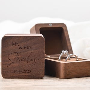 May include: A wooden ring box with the inscription "Mr. & Mrs. Sherley 10.16.2025" engraved on the lid. The box is open, revealing two rings. The box is made of dark wood.