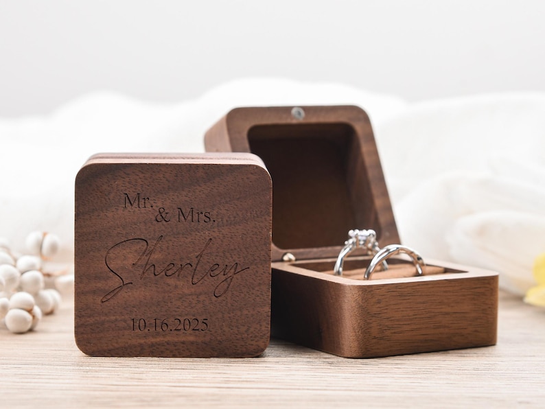 May include: A wooden ring box with the inscription "Mr. & Mrs. Sherley 10.16.2025" engraved on the lid. The box is open, revealing two rings. The box is made of dark wood.
