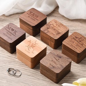 May include: A collection of personalized wooden ring boxes. Each box is square with a hinged lid and features engraved names, initials, and dates. The boxes are made of various wood tones, including dark walnut and lighter wood, and are arranged on a light wooden surface.