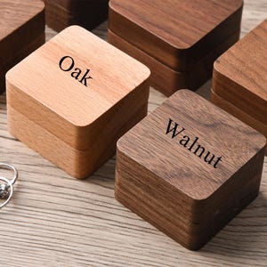 May include: Wooden ring boxes in various shades of brown, including oak and walnut. The boxes are square with a hinged lid. The word "Oak" is printed on one box, and "Walnut" is printed on another. Two silver rings are in the foreground.