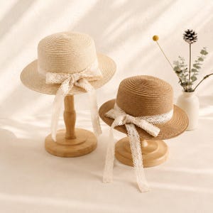 May include: Two straw hats with flat crowns and wide brims, each featuring a white lace ribbon tied in a bow around the base of the crown. The hats are displayed on wooden stands, with one hat in a light beige tone and the other in a darker tan.