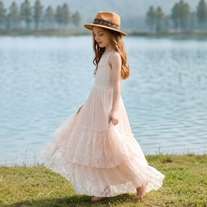 Puede incluir: Un vestido largo sin mangas de color rosa claro con falda de niveles con ribete de encaje y un sombrero fedora a juego con una banda estampada. El vestido tiene un corpiño ajustado con detalles de encaje en el cuello y una falda plisada.