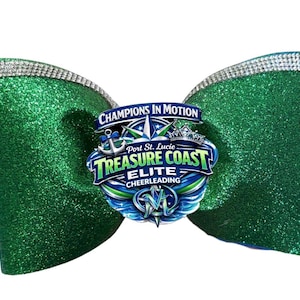 May include: A large green glitter cheerleading bow with a silver rhinestone trim. The bow features a logo that reads "Champions in Motion" and "Treasure Coast Elite Cheerleading" in blue, green, and white.