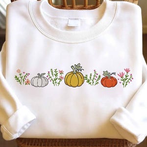May include: White sweatshirt featuring a row of three pumpkins in gray, yellow, and orange, with green vines and pink flowers. The design is centered across the chest. The sweatshirt has long sleeves and a crew neck.