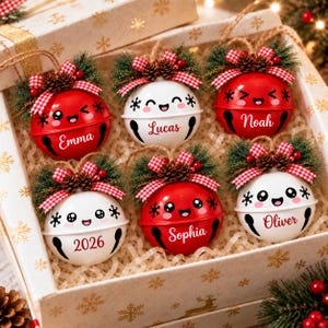 May include: A set of six round jingle bell ornaments in red and white, each featuring a cartoon face, a name or year, and a decorative topper with pine needles, small pinecones, red berries, and a red and white checkered ribbon bow.
