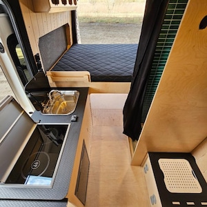 Puede incluir: Vista interior de una furgoneta camperizada con cocina, cama y almacenamiento integrados. La cocina cuenta con un fregadero y una placa de cocina. La cama tiene un colchón acolchado negro. Las paredes y los armarios son de madera clara.