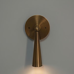 May include: A brass wall sconce with a circular backplate and a conical shade. The sconce emits a warm, downward-facing light, creating a focused illumination. The fixture is mounted on a light gray wall.