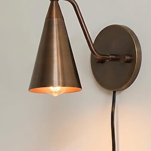 May include: A wall-mounted sconce lamp with a conical bronze-toned metal shade and a matching circular wall plate. The fixture features an adjustable arm and a black power cord that plugs into a standard wall outlet, emitting a warm light.