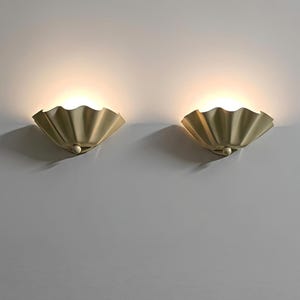 Peut inclure: Une paire d'appliques murales au fini doré mat, présentant une forme de coquille plissée en éventail. Chaque luminaire diffuse une lueur chaude et douce par le haut, projetant une lumière délicate vers le haut contre la surface du mur.